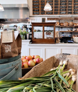 A cozy bakery counter with loaves of bread, apples, and fresh green onions on display. A chalkboard menu with drinks hangs above, and pendant lights illuminate the bright, rustic interior.