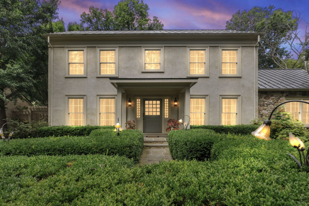 A two-story house with beige walls, shuttered windows, and a central front door stands amidst neatly trimmed hedges and garden lights at dusk—radiating balance and harmony reminiscent of Libra season.