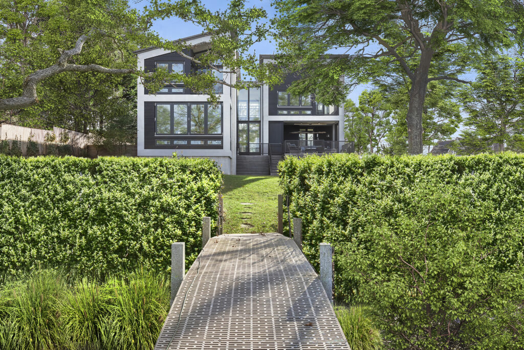 A modern house with large glass windows sits behind lush green hedges and trees, viewed from a metal dock leading up from the water through the garden—an inviting retreat perfect for relaxing during libra season.