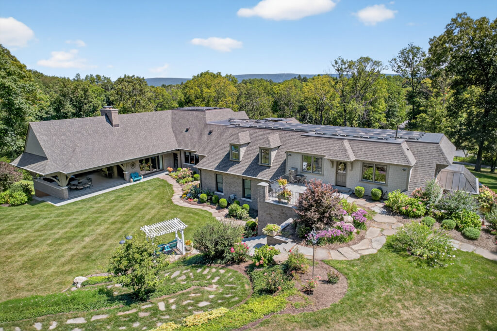 A large house with a gray roof, solar panels, landscaped gardens, stone pathways, a carport, and a pergola sits among trees and green lawns under a blue sky with scattered clouds.