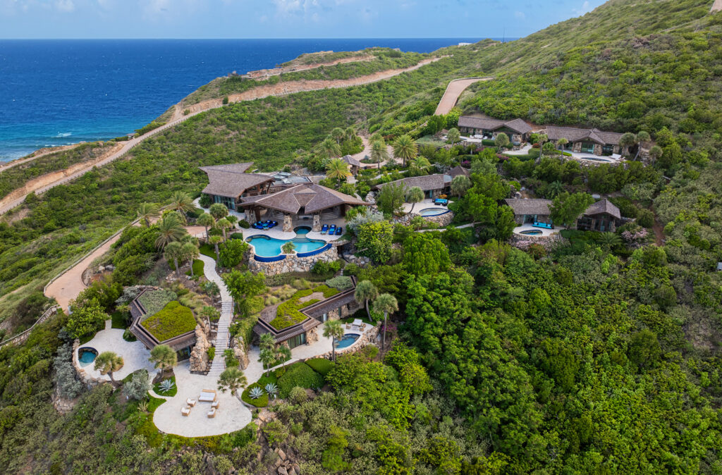Aerial view of a luxurious hillside estate with multiple swimming pools, thatched-roof buildings, blue lounge chairs, and ocean views surrounded by lush greenery.