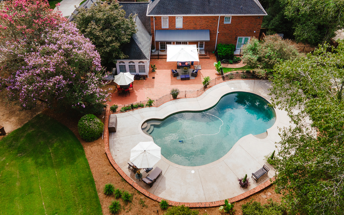 Aerial view of a backyard oasis in a vibrant college town, featuring a kidney-shaped swimming pool, lounge chairs, umbrellas, patio seating, and lush landscaping beside a brick house.