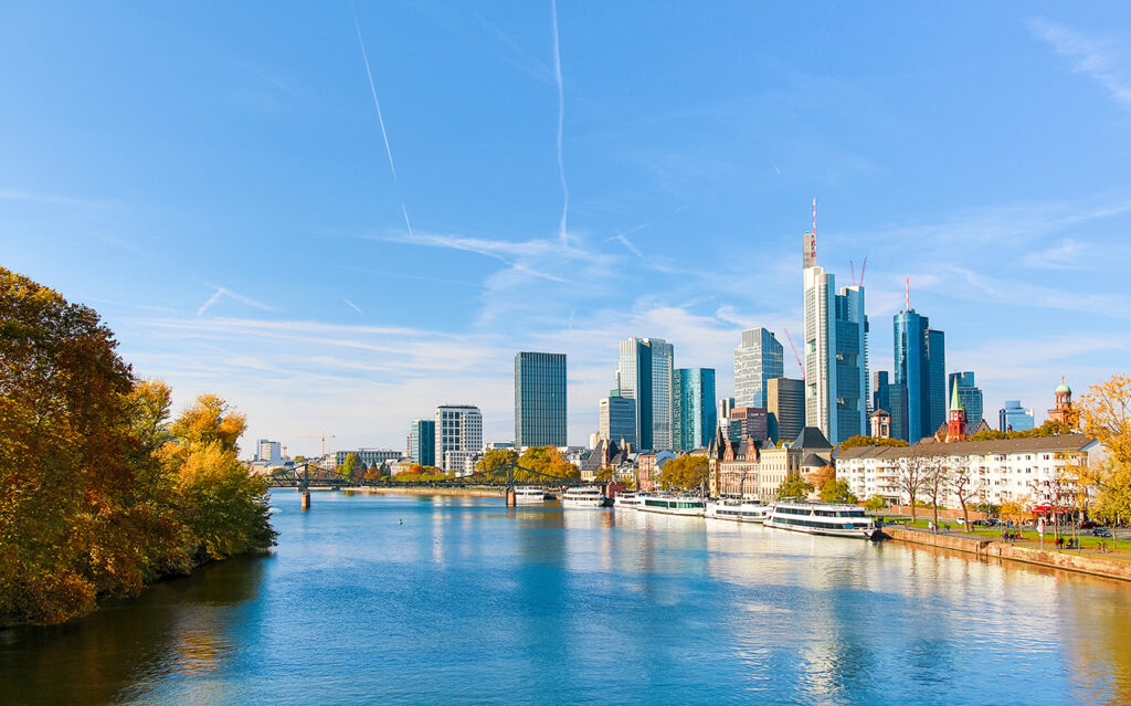 frankfurt_2 A wide river flows through a city with modern skyscrapers and tall glass buildings under a clear blue sky. Trees with autumn foliage line the riverbank, and boats are docked along the shore.