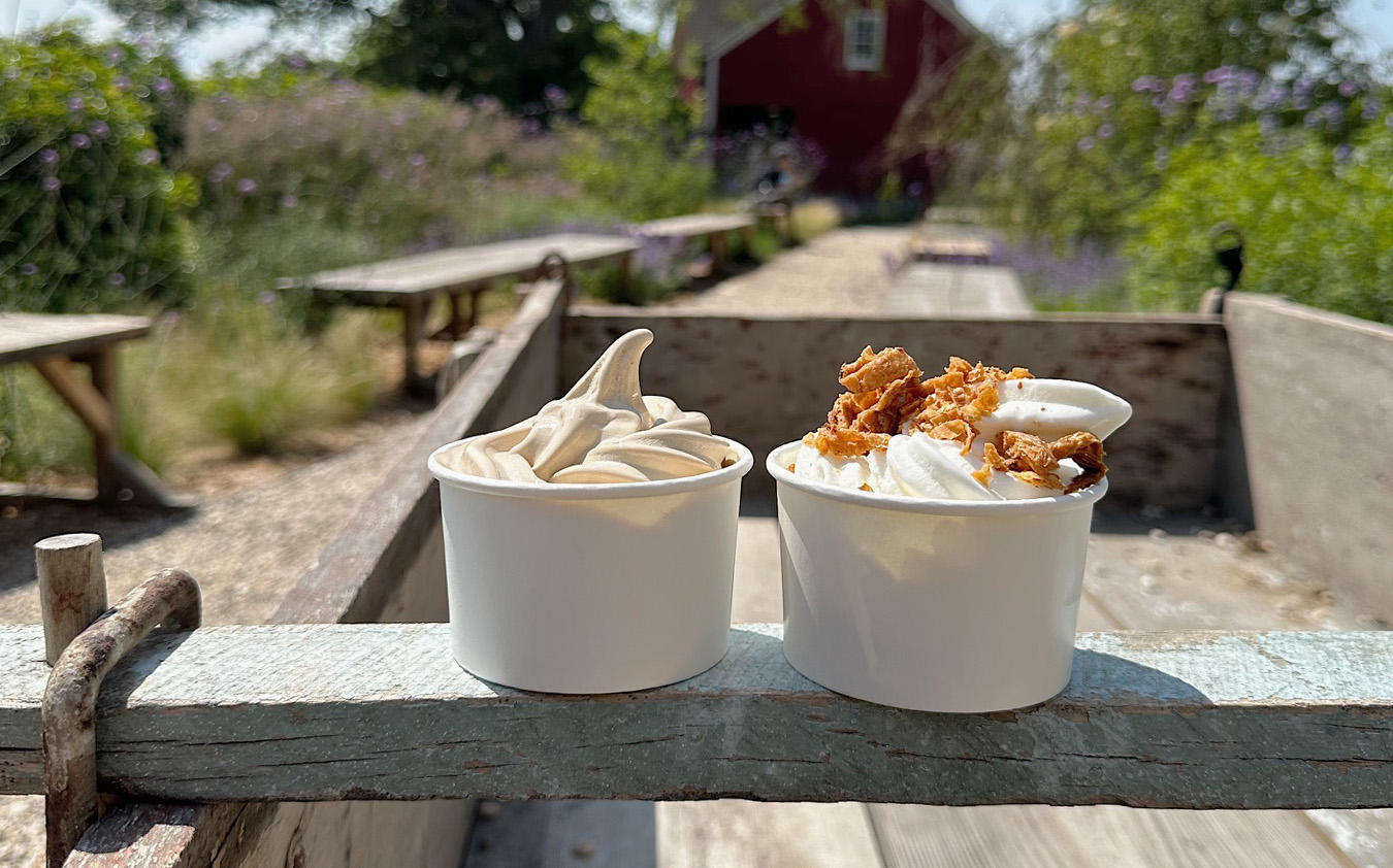 Two cups of soft-serve ice cream, one plain and one topped with crumbled pieces, sit on the edge of a rustic wooden cart outdoors, with a blurred garden and red barn in the background.