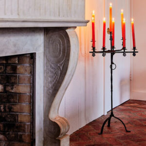 A black metal candelabrum with seven lit red candles stands on a brick floor beside a white marble fireplace with ornate carving, while vintage cookbooks rest nearby.