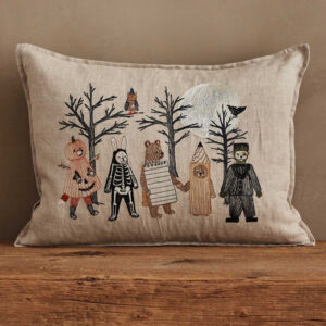 A linen pillow features embroidered animals and figures in Halloween costumes—like a pumpkin, skeleton, pencil, and suit—standing before leafless trees with a full moon, an owl, a bat, and stacked cookbooks in the background.