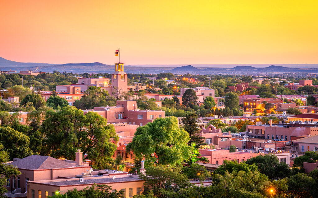View of Santa Fe, New Mexico at sunset, with adobe-style buildings, lush green trees, and the New Mexico State Capitol building featuring a flag, set against a glowing yellow-orange sky.