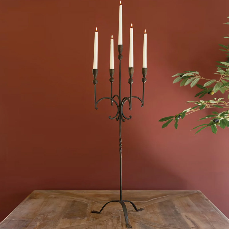 A tall black metal candelabra with five lit white taper candles stands on a wooden table against a red wall, surrounded by an inviting stack of cookbooks, with green plant leaves visible on the right side.