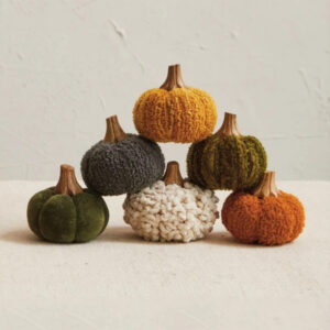 Six decorative pumpkins made of textured fabric in green, yellow, orange, cream, and gray colors are arranged in a pyramid on a light, textured surface against a plain wall—perfect for adding autumn charm to your kitchen shelf among your favorite cookbooks.