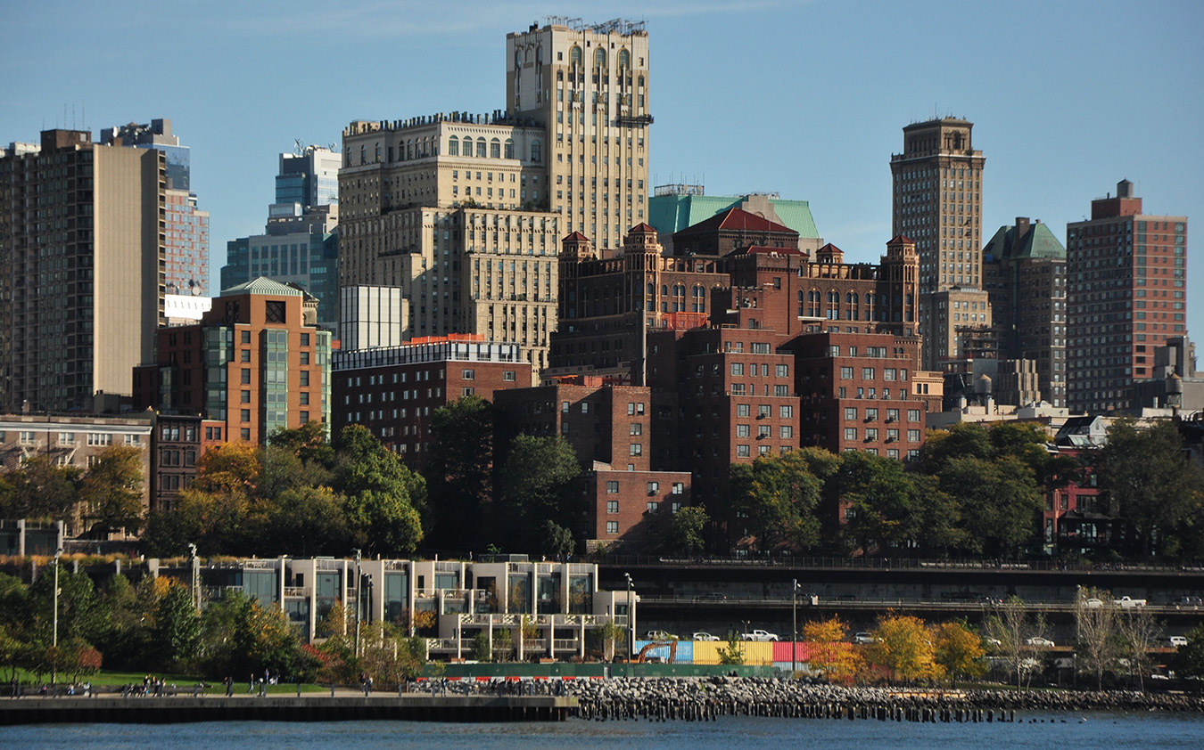A cityscape view of Brooklyn Heights, New York, showcases various tall nyc residential rental and office buildings, trees along the waterfront, and a clear blue sky in the background.