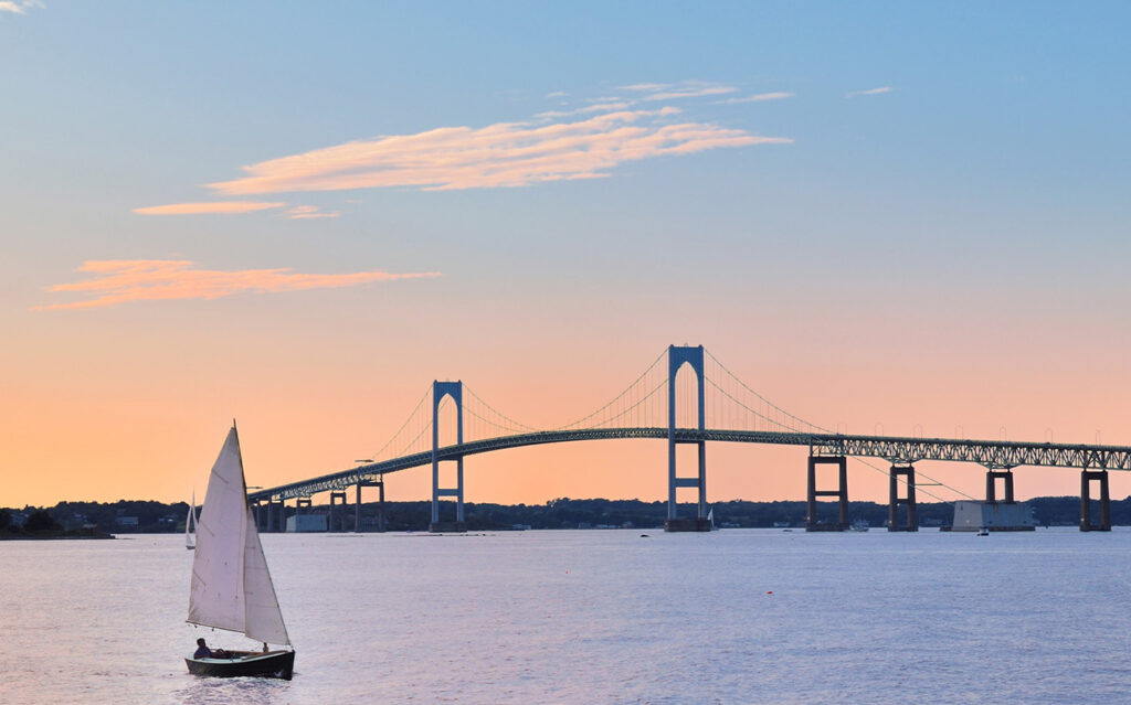 A small sailboat glides on calm water at sunset, with a large suspension bridge spanning the background under a pastel sky.