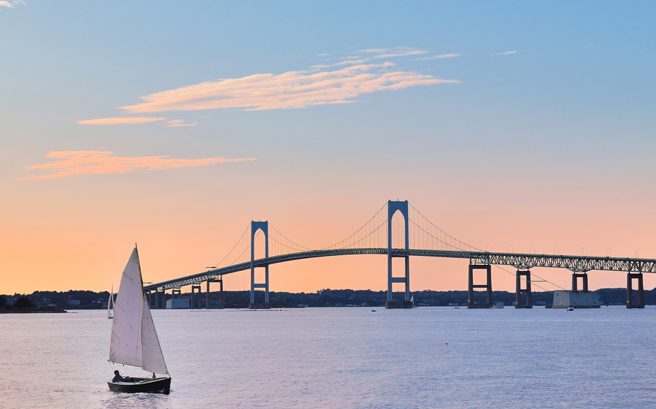 A small sailboat glides on calm water at sunset, with a large suspension bridge spanning the background under a pastel sky.