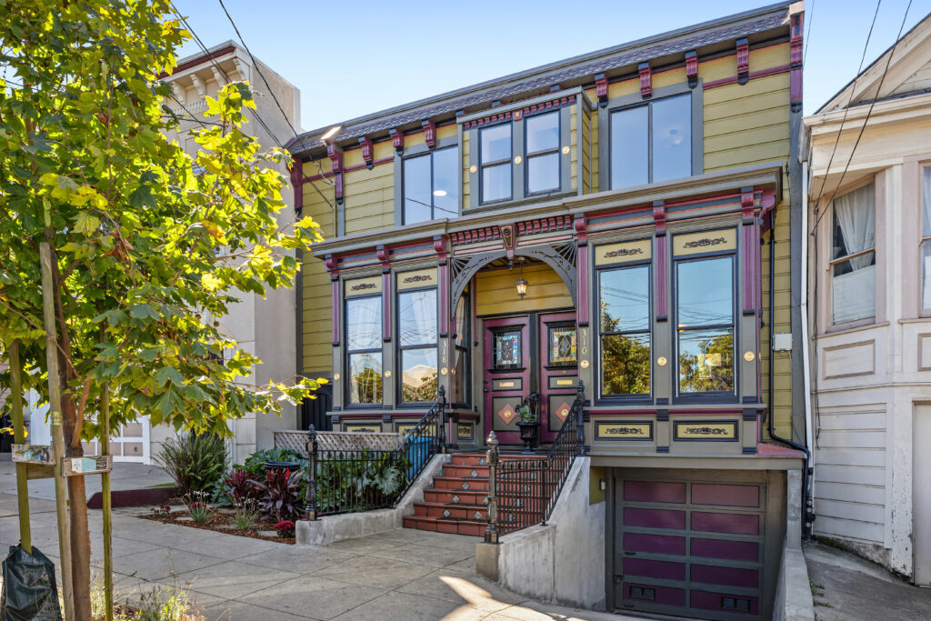 A two-story Victorian house with green and burgundy trim, large front windows, ornate detailing, and a red stairway leading to a double front door. Reminiscent of Caribbean houses, a garage sits at street level, and a tree stands on the sidewalk.