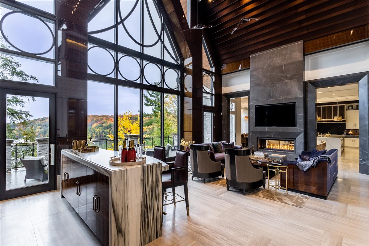A modern, open-concept living space features a marble kitchen island, dark wood accents, a fireplace with a mounted TV, floor-to-ceiling windows, and a scenic view of trees in autumn.