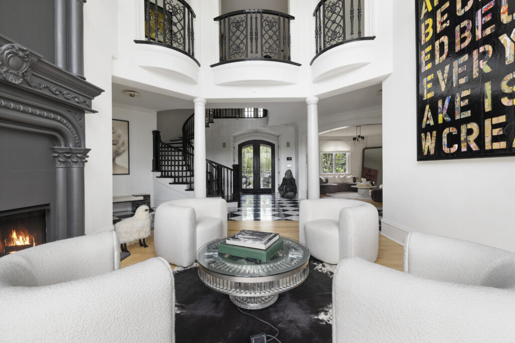 2410 Hidden River Lane Franklin, TN 37069 A modern living room with four white chairs around a glass coffee table, a black fireplace, abstract wall art, a spiral staircase, and a balcony overlooking the space.