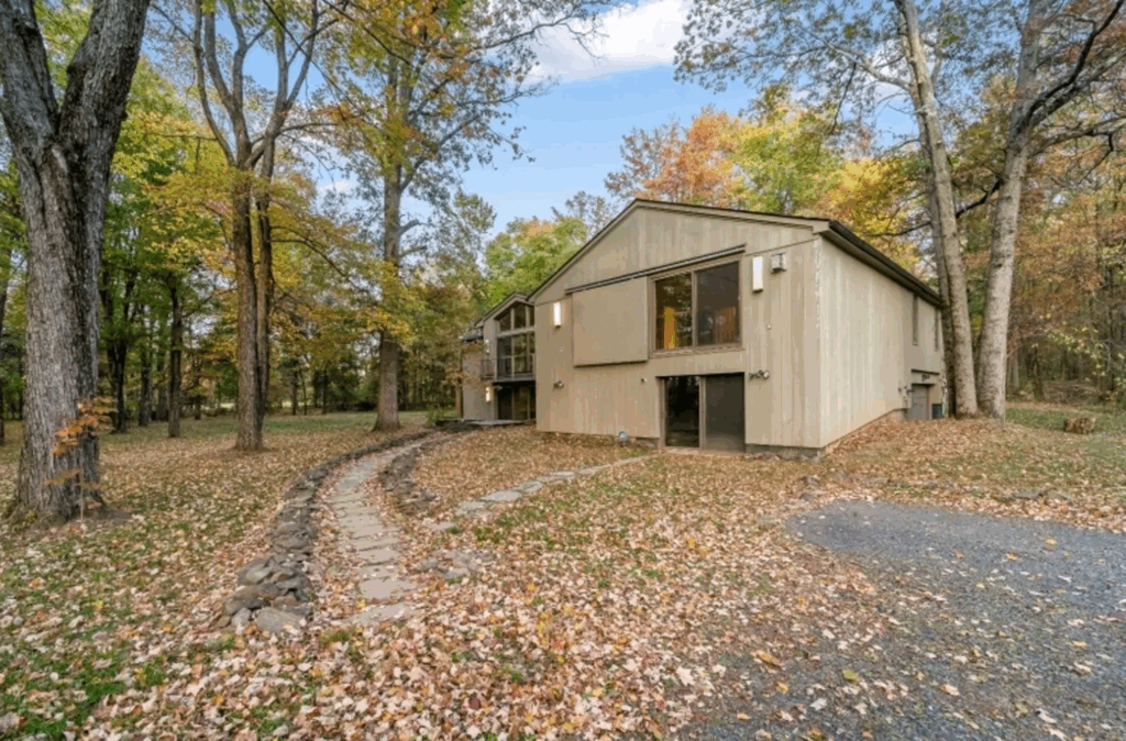 288 County Road 519 A modern, two-story house with large windows sits among tall trees on a property covered with vibrant fall foliage. A stone path leads from the driveway to the entrance, surrounded by a wooded landscape.