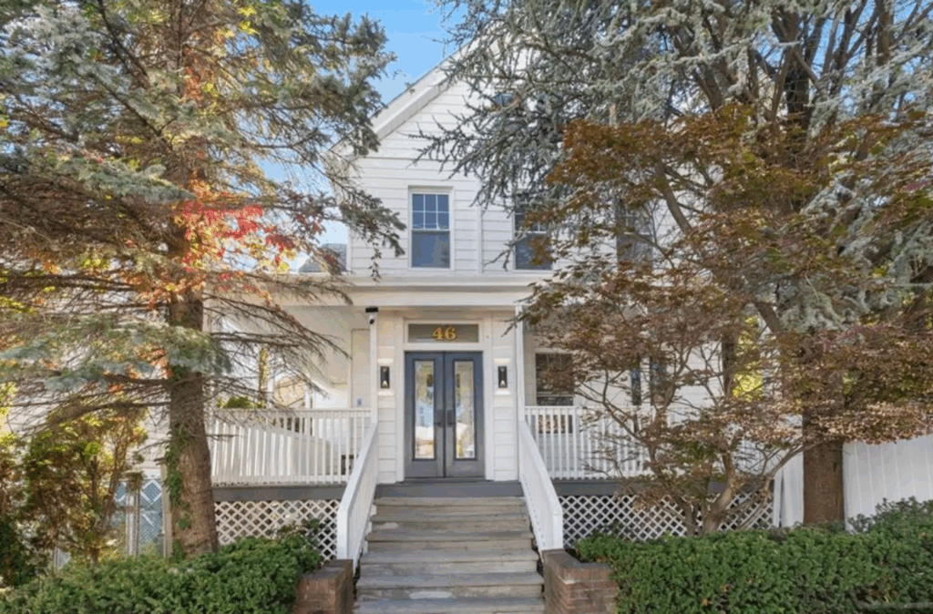 46 Garden Place A white two-story house with a wide front porch, stairs leading up, double front doors, and large trees adorned in fall foliage partially framing the entrance.