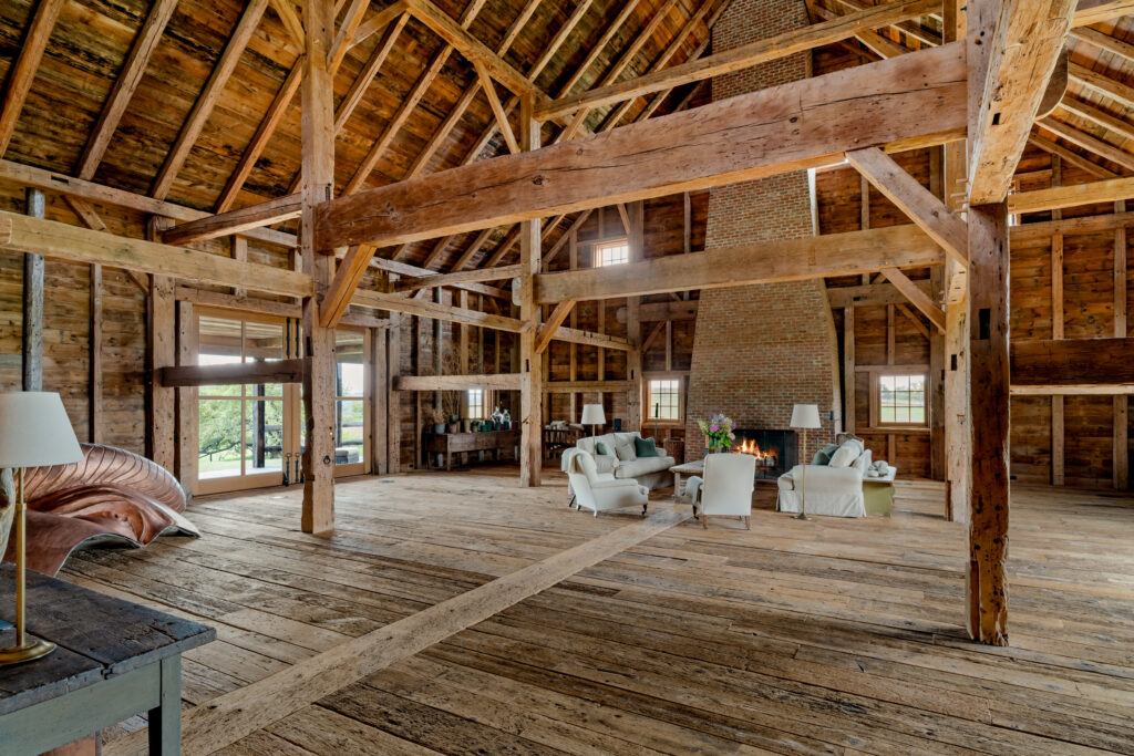 118 Finkle Road ANCRAMDALE, NY 12503 A spacious, rustic room with exposed wooden beams, high ceilings, and a brick fireplace. White armchairs and a couch sit near the fire, and large windows let in natural light, revealing views of greenery outside.