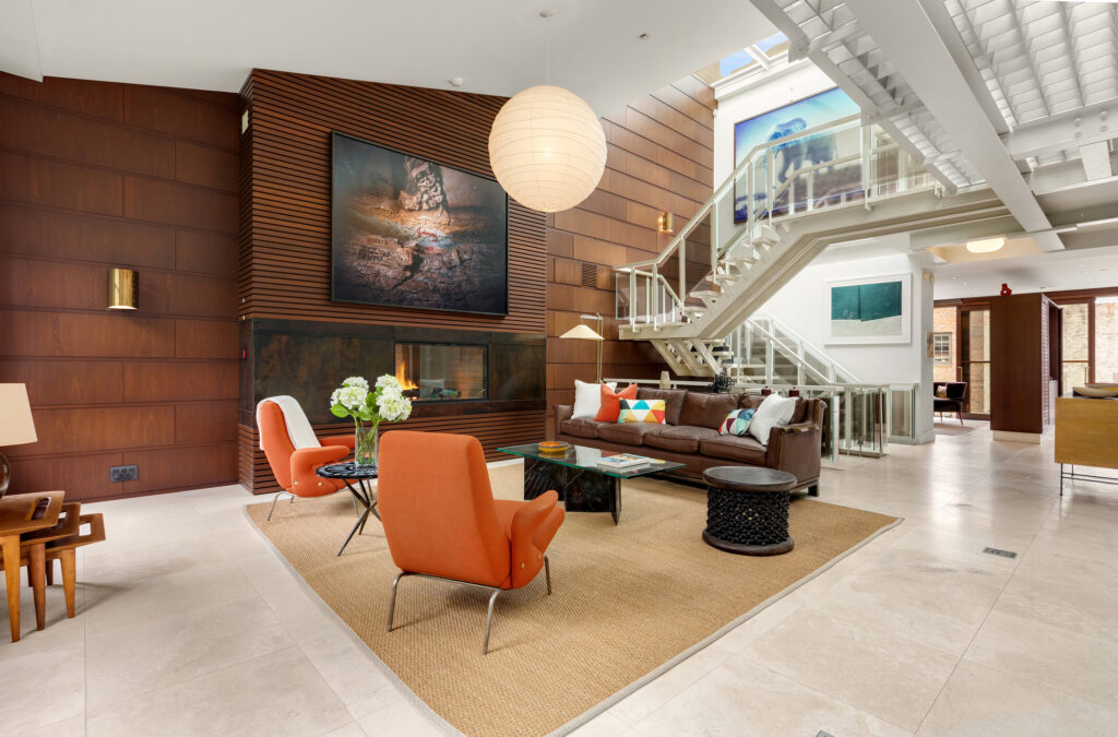 22 Gramercy Park South, PH Gramercy Park, Manhattan, Modern living room with two orange chairs, a brown sofa, a glass coffee table, large wall art, a fireplace, and a floating staircase with glass railing. Natural light fills the space through large windows and skylights.