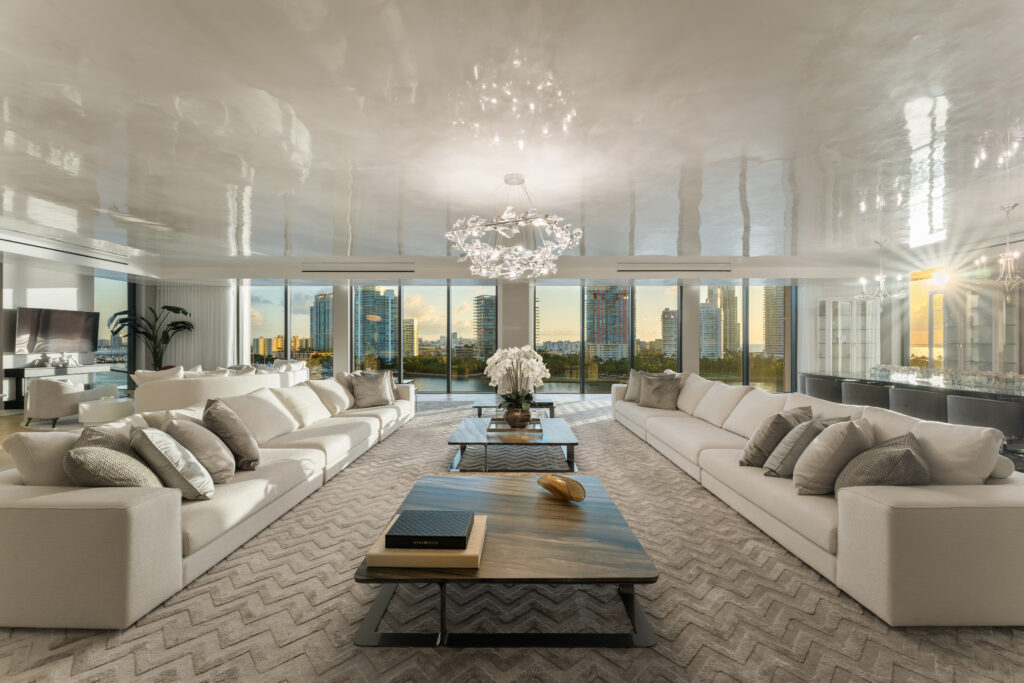 6893 Fisher Island Dr, LPH 6893 Miami Beach, FL Luxurious living room with white sofas, glass coffee tables, a large chandelier, and floor-to-ceiling windows offering a city skyline and sunset view. The space is modern, bright, and elegantly decorated.
