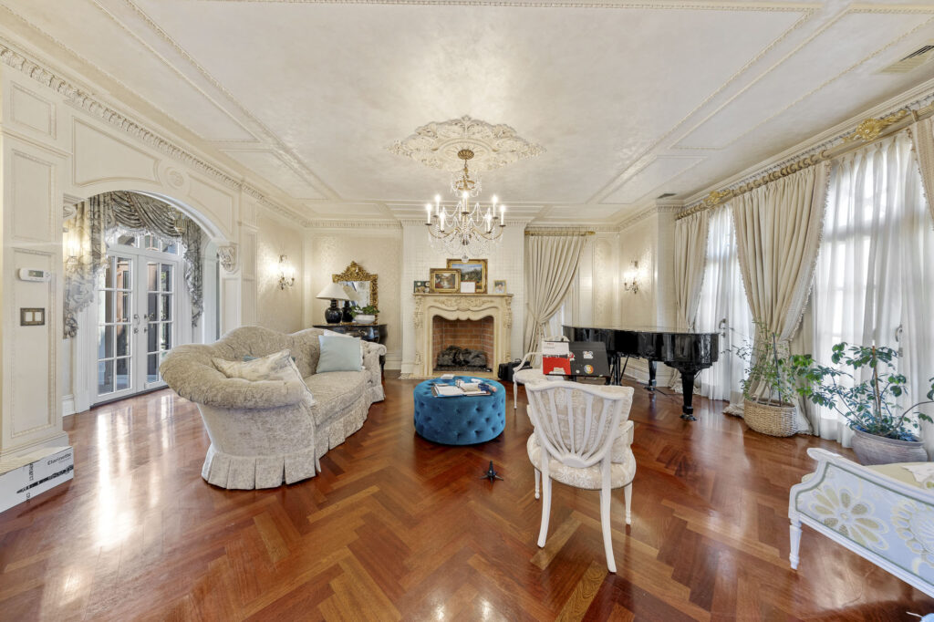 78 Roberts Road Englewood Cliffs, NJ 07632 Elegant living room with ornate molding, chandelier, large fireplace, grand piano, plush sofas, blue ottoman, and parquet wood floor. Tall windows with long drapes let in natural light; plants and artwork add detail.