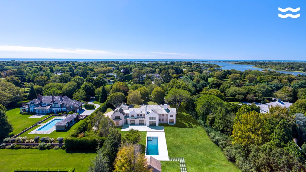 Aerial view of large estates with swimming pools nestled among green trees and lawns, with a lake and ocean in the background under a clear blue sky—perfect for reflecting on astrological houses during scorpio season.