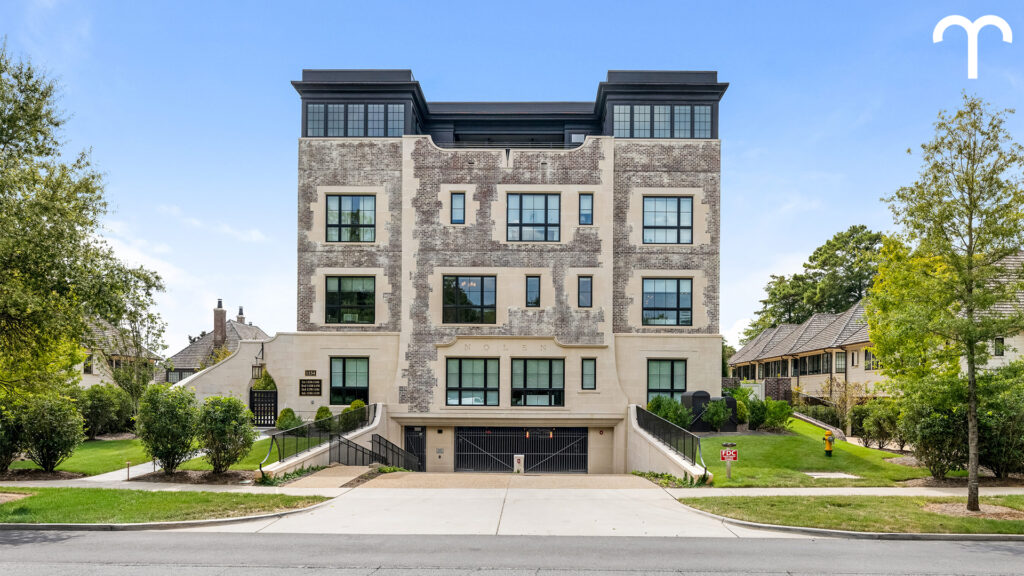 A modern four-story apartment building with large windows, a faded brick exterior, and dual driveways leading to an underground garage, surrounded by green lawns and neighboring houses.