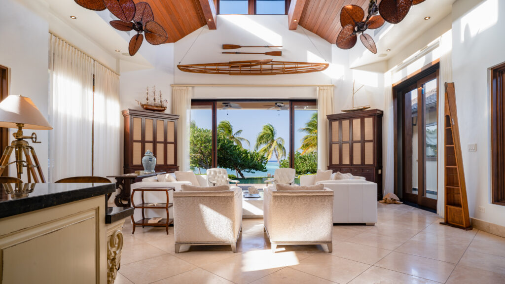 Bella Beach, Oil Nut Bay Virgin Gorda A bright, airy living room with white furniture, wood accents, and large glass doors opening to palm trees and an ocean view, with natural light streaming from skylights above.