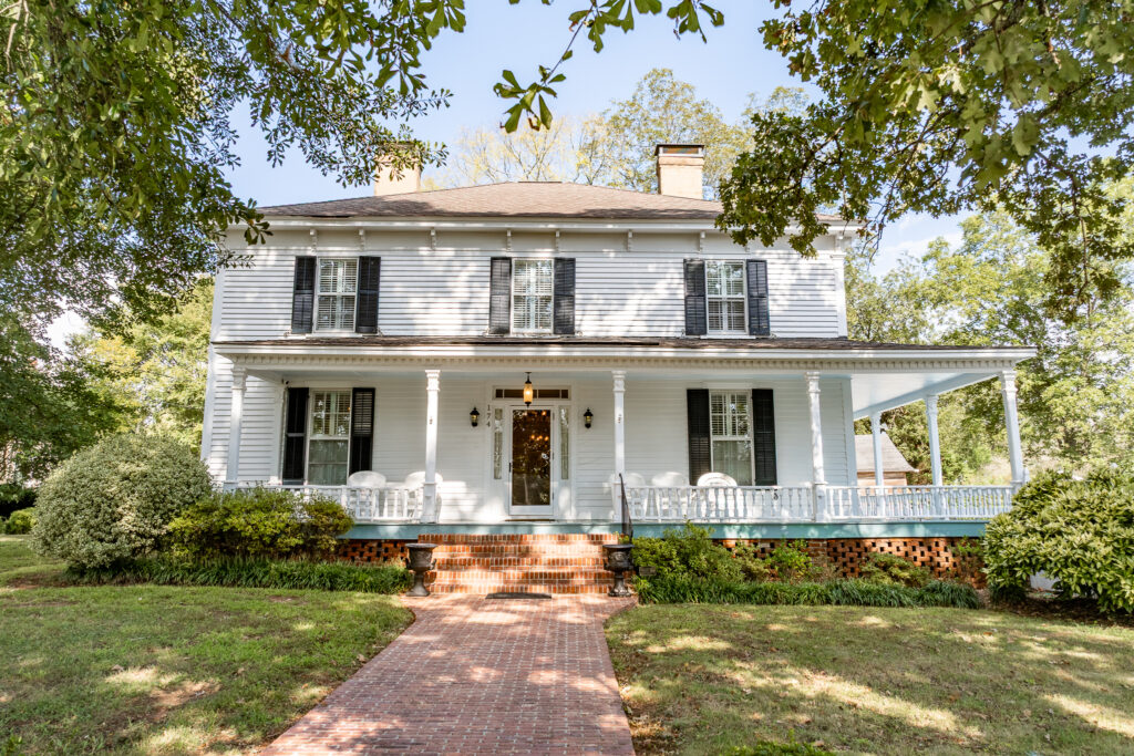 A two-story white house with black shutters, a wide front porch, and brick steps recalls the charm of Caribbean houses, surrounded by green bushes and trees under a clear blue sky.