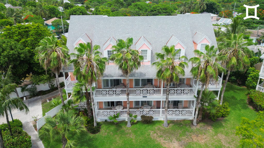 A three-story building with pink and white accents, surrounded by palm trees and greenery, stands under a partly cloudy sky.