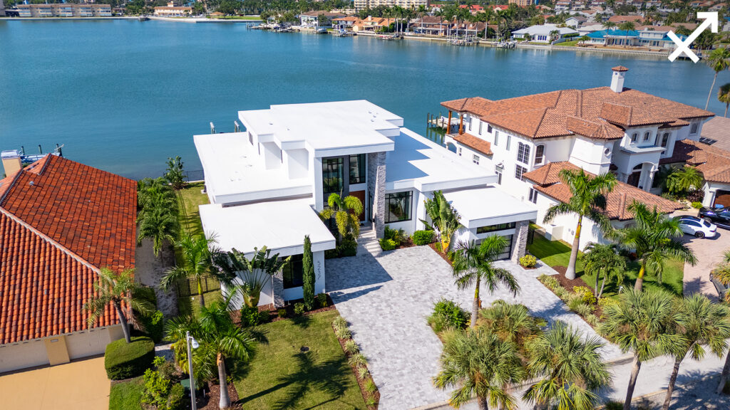 Aerial view of a modern white waterfront house with a flat roof, palm trees, and a dock, set beside similar upscale homes, overlooking a calm bay—perfect for reflection during Scorpio season. An arrow icon appears in the top right corner.