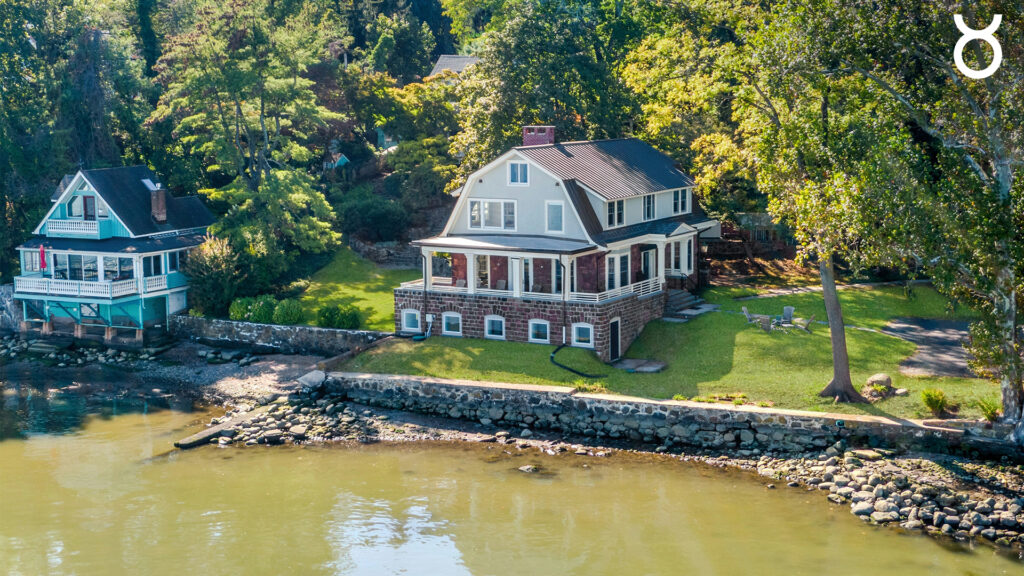 A large, two-story house with a screened porch and stone foundation sits by the water, surrounded by trees and a grassy yard. A stone wall lines the shoreline next to a smaller blue house.
