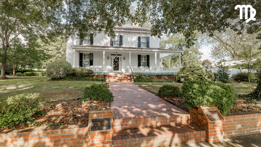 A two-story white house with green shutters and a front porch sits behind a brick walkway and low brick wall, surrounded by trees and well-kept bushes.