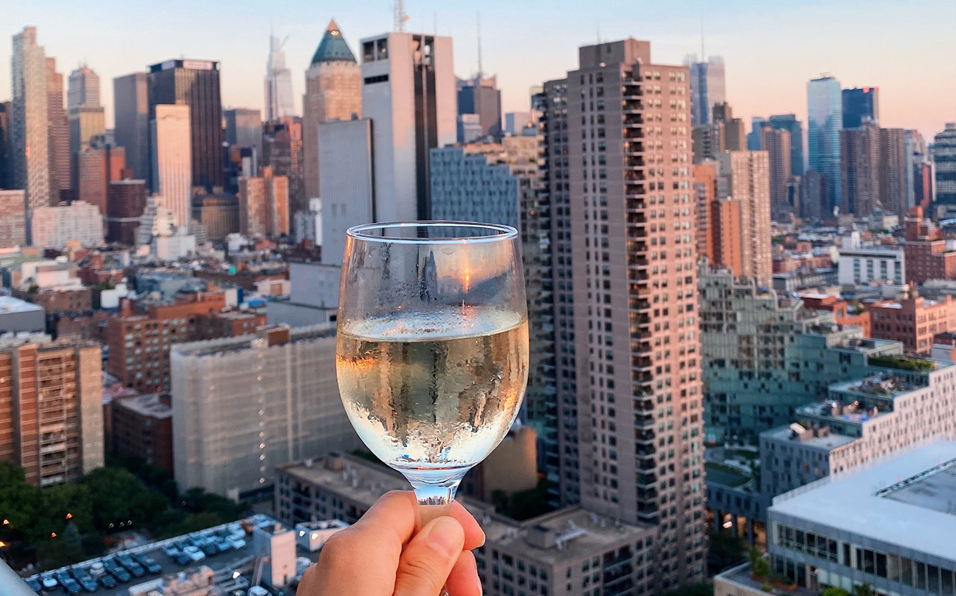 A hand holds a glass of white wine up in front of a city skyline with tall buildings at sunset, with the glass in focus and the cityscape blurred in the background.