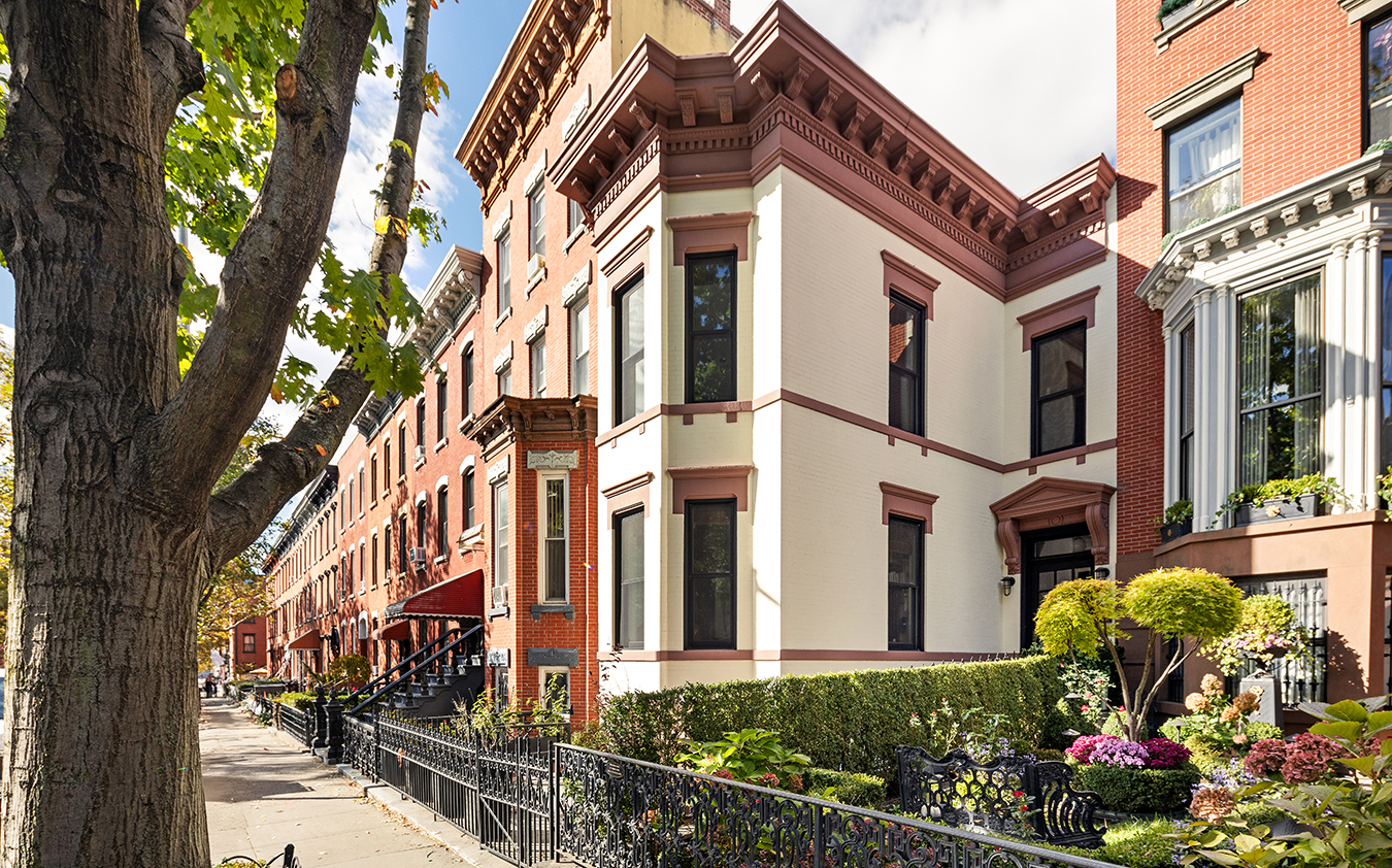 A row of historic townhouse brownstones with decorative trim and railings lines a sunny, tree-shaded street, featuring lush gardens in front.
