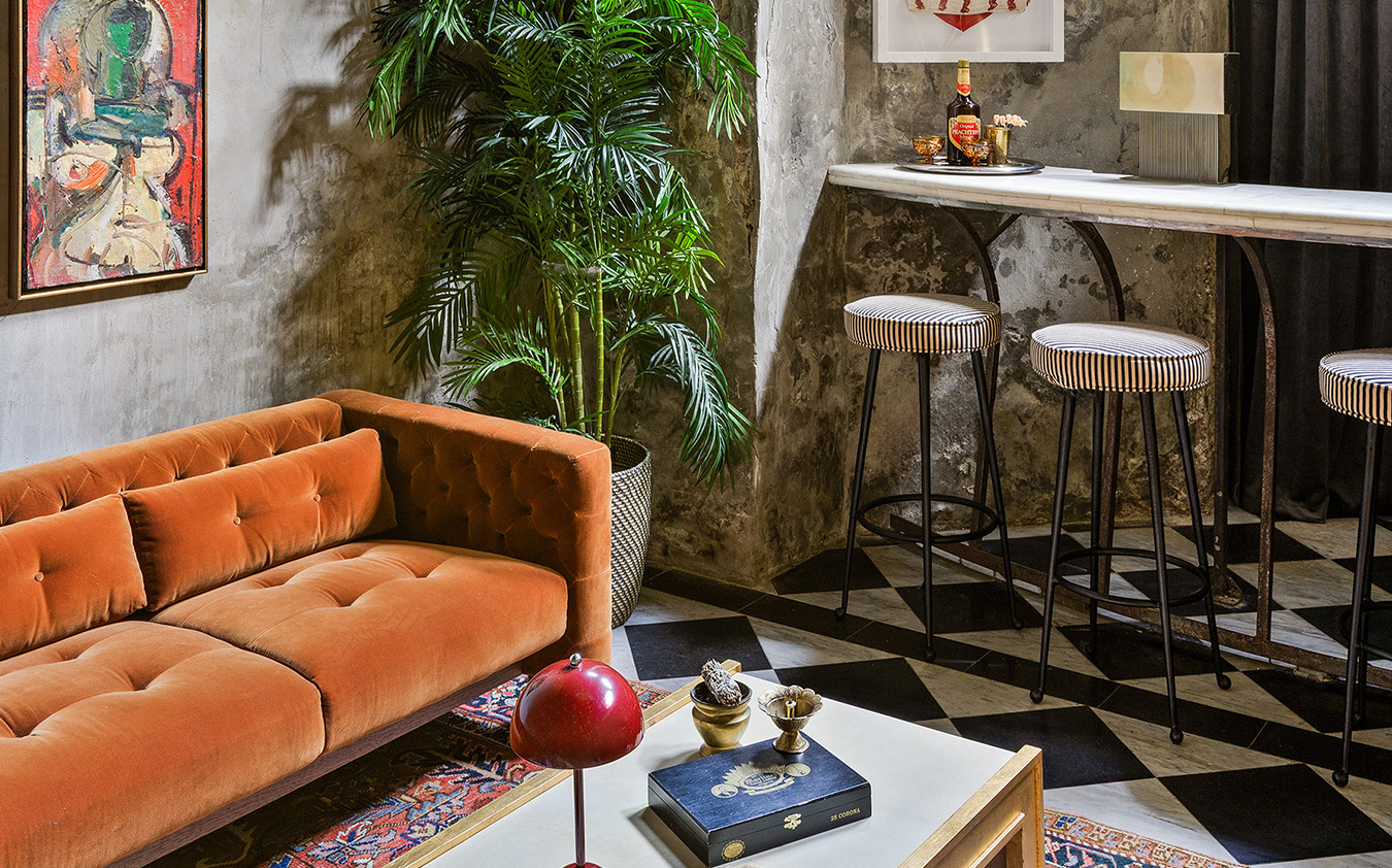 A cozy living area with an orange tufted sofa, a coffee table with decor, a potted plant, and abstract art sets the scene for an A-list November 2025 gathering by the marble-topped bar and striped stools atop the checkered floor.