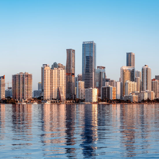 A city skyline with modern high-rise buildings reflecting on calm water under a clear blue sky, illuminated by warm sunlight.