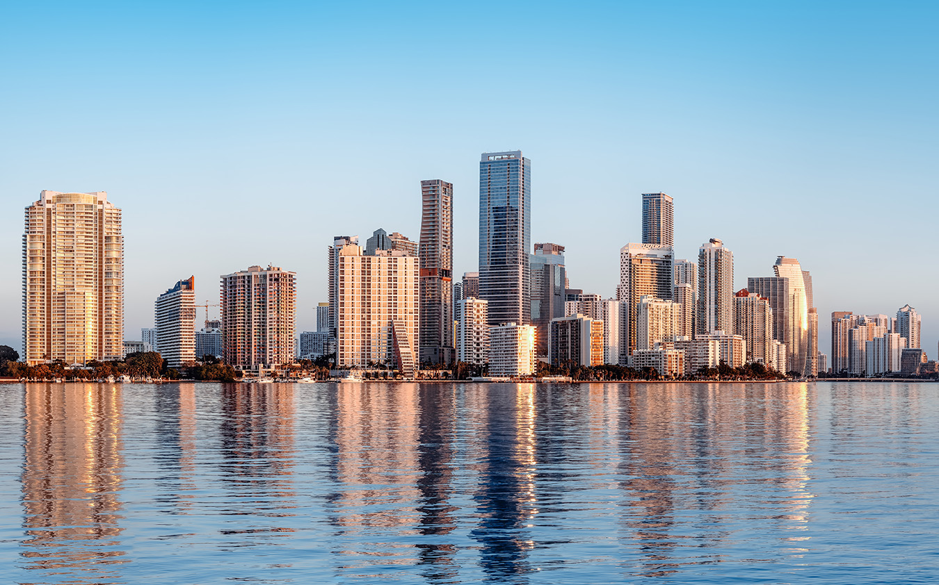 A city skyline with modern high-rise buildings reflecting on calm water under a clear blue sky, illuminated by warm sunlight.