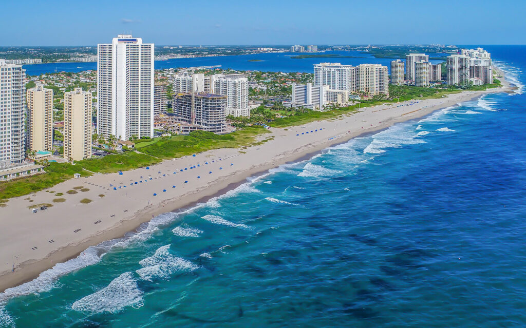 Aerial view of a sandy beach lined with high-rise buildings along the shoreline. Gentle waves roll in from the vibrant blue ocean, and umbrellas are spaced along the beach. Greenery and more buildings are visible in the background.