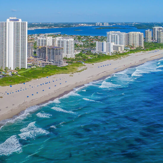 Aerial view of a sandy beach lined with high-rise buildings along the shoreline. Gentle waves roll in from the vibrant blue ocean, and umbrellas are spaced along the beach. Greenery and more buildings are visible in the background.