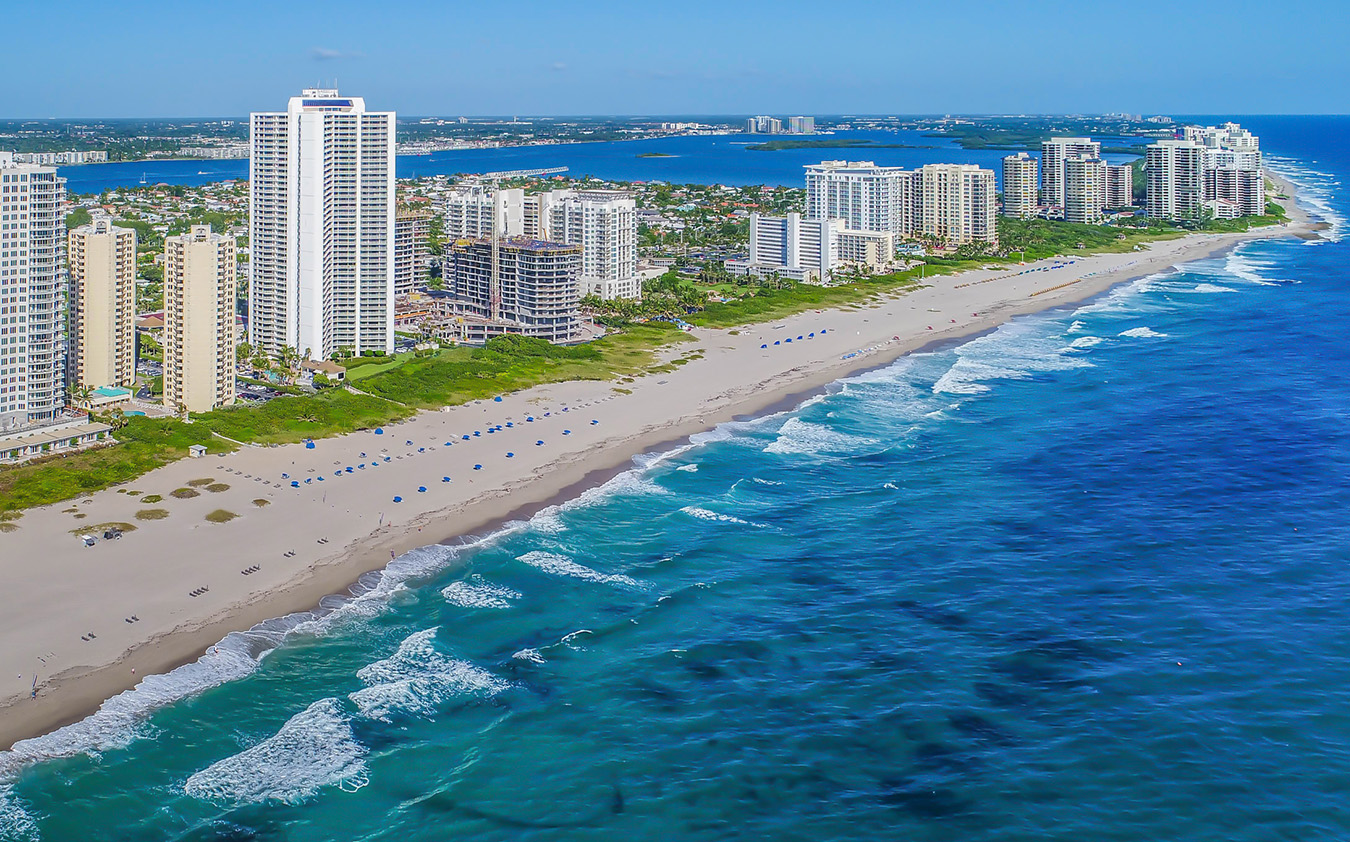 Aerial view of a sandy beach lined with high-rise buildings along the shoreline. Gentle waves roll in from the vibrant blue ocean, and umbrellas are spaced along the beach. Greenery and more buildings are visible in the background.