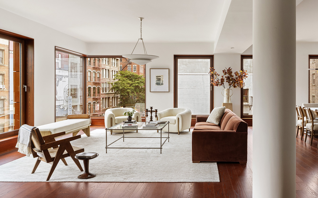 A modern, sunlit living room with large windows, a beige rug, a glass coffee table, cream armchairs, a brown velvet sofa, and wooden floors. Decor includes books, vases, and wall art, with a dining area visible in the background.