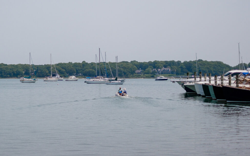 th_eastend A small motorboat moves away from a dock lined with yachts on a calm bay, with several sailboats anchored nearby—an inviting scene that reflects the tranquil lifestyle offered by East End Real Estate. Trees and a hazy sky complete the picturesque backdrop.