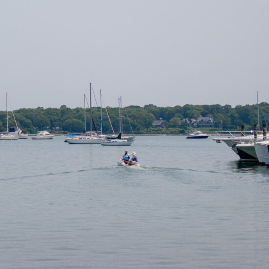 A small motorboat moves away from a dock lined with yachts on a calm bay, with several sailboats anchored nearby—an inviting scene that reflects the tranquil lifestyle offered by East End Real Estate. Trees and a hazy sky complete the picturesque backdrop.