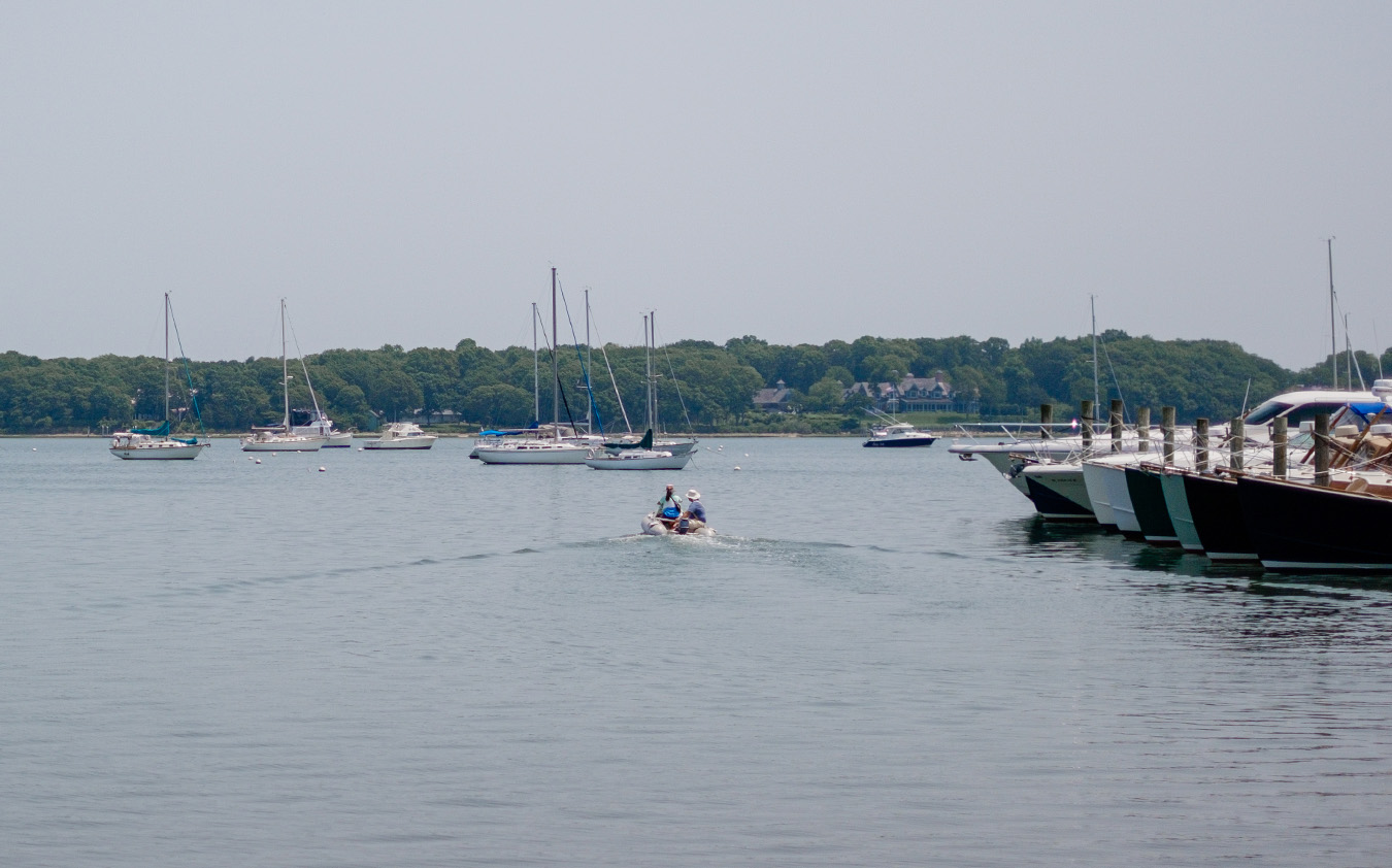 A small motorboat moves away from a dock lined with yachts on a calm bay, with several sailboats anchored nearby—an inviting scene that reflects the tranquil lifestyle offered by East End Real Estate. Trees and a hazy sky complete the picturesque backdrop.