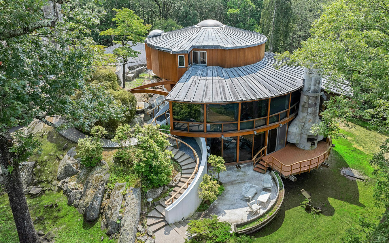 Aerial view of a modern, circular house with large glass windows, curved walls, and a metal roof, surrounded by trees, rocks, and greenery. Several patios and stairways integrate the home with the natural landscape.