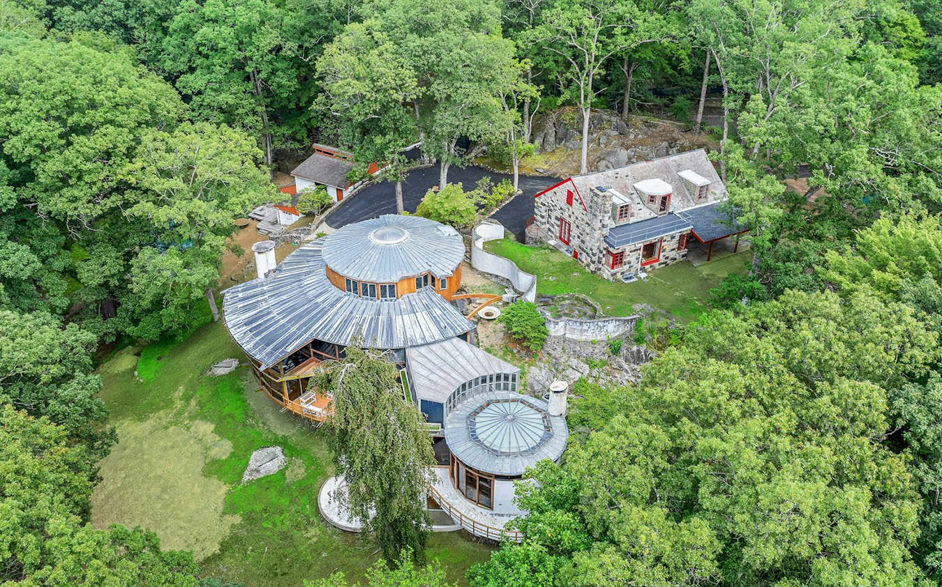 th_gleason Aerial view of two unique homes surrounded by dense green trees, featuring circular and irregular roof designs, stone walls, and pathways, with a driveway winding through the wooded area.