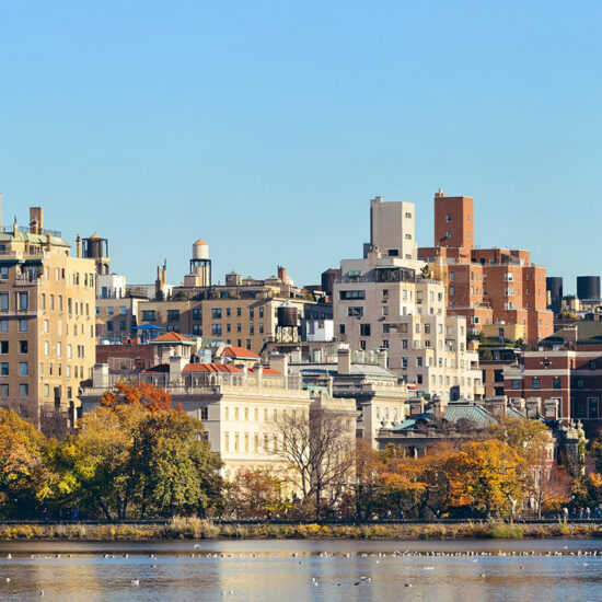 A cityscape showing mid-rise buildings with varied architecture behind colorful autumn trees, all reflected in a calm body of water under a clear blue sky—capturing the elegance often seen in Manhattan luxury sales.