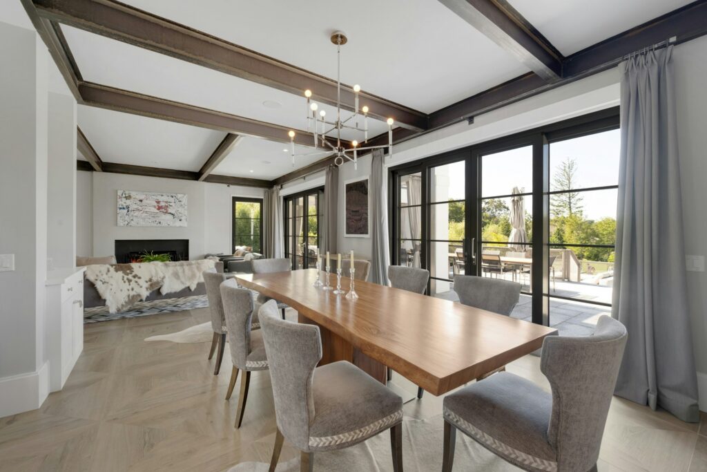 Modern dining rooms shine with a wooden table, six gray upholstered chairs, large windows with gray curtains, and a chandelier. The space seamlessly opens to a living area featuring a sofa and stylish cowhide rug.
