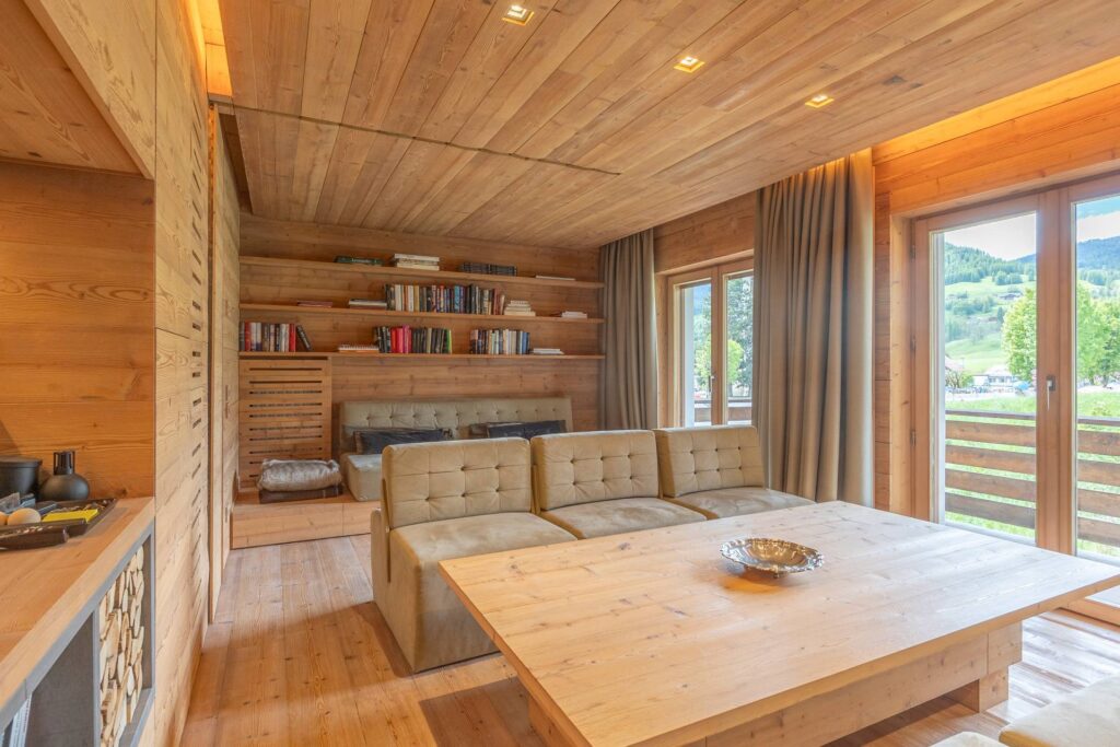 A cozy wooden living room, with beige sofas, a large wooden table, bookshelves, and big windows framed by curtains, offering views of green hills. Warm lighting highlights the natural wood interior.